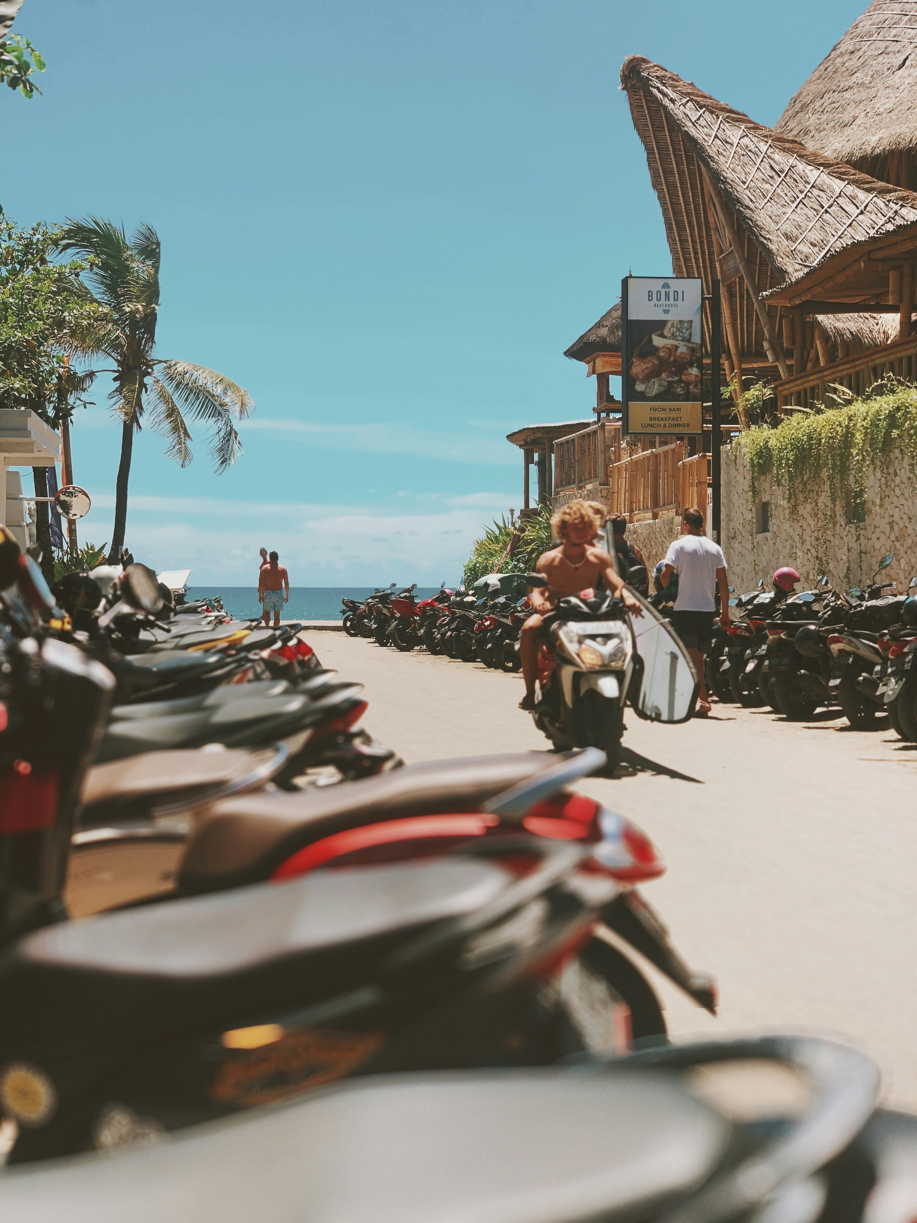 Scooters lined along a sunlit street in Canggu, with surfers, palm trees, and beachside caf&eacute;s leading down towards the sea, capturing the energy and congestion of Bali&rsquo;s coastal hotspots.